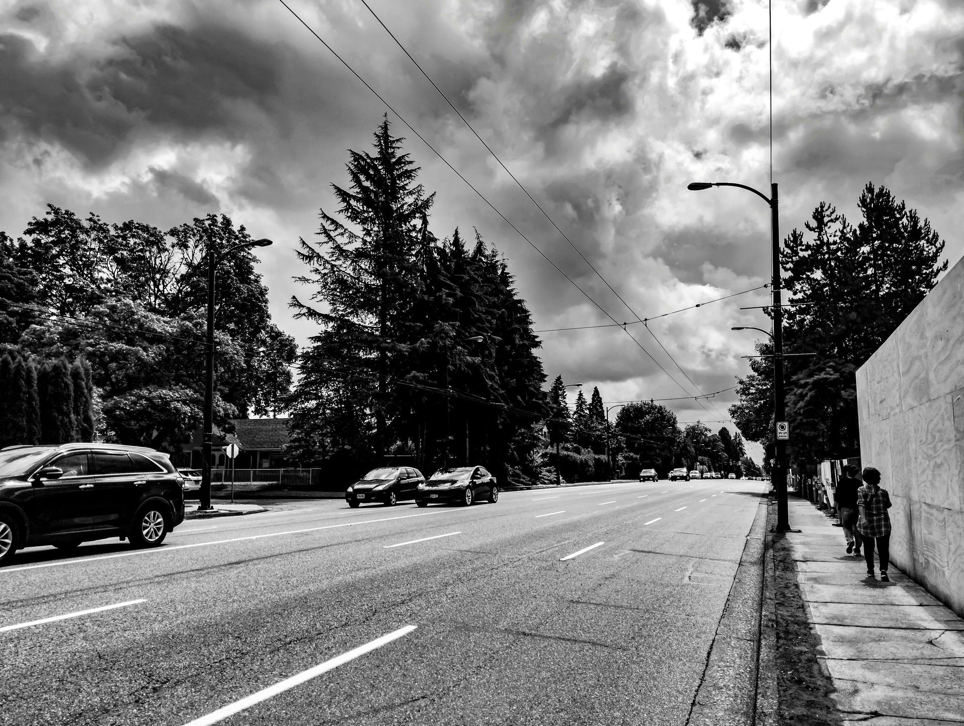 Black and white street view with cars, trees, cloudy sky, and pedestrians on the sidewalk.
