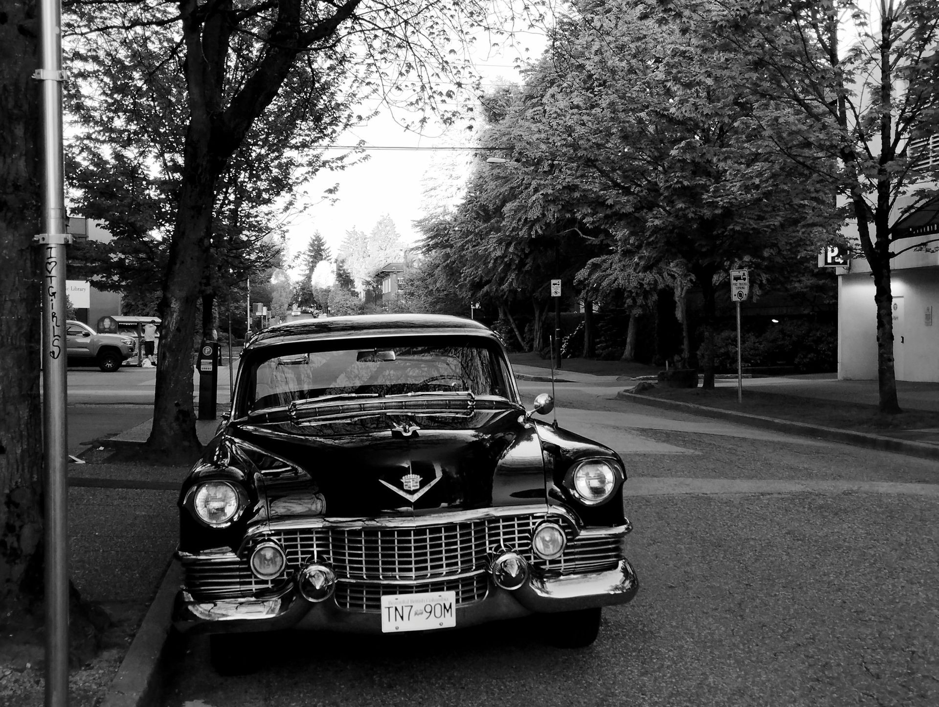 Black and white image of a vintage car parked on a quiet street lined with trees and buildings.